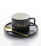 Black ceramic cup with 'Best wishes' gold text, matching saucer, and gold spoon on white background